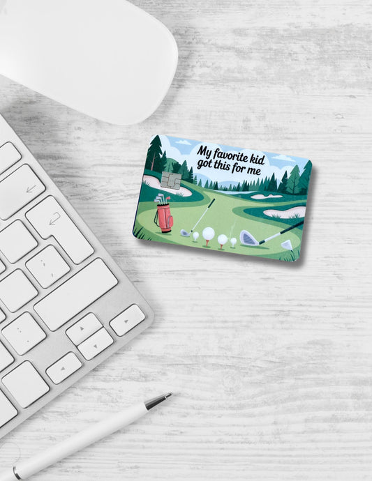 A golf-themed debit card skin sits on a white wood desk next to a wireless mouse, computer keyboard, and pen. The card design features a green golf course, golf clubs, balls, and the humorous phrase &quot;My favorite kid got this for me.&quot; It&#39;s a playful, sporty accessory with a lighthearted gift message perfect for Father&#39;s Day.