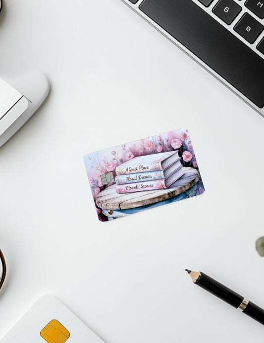A pastel floral-themed debit card skin lies on a white desk beside a laptop, pen, and smart card reader. The card features a romantic book stack with titles &quot;A Quiet Place,&quot; &quot;Floral Dreams,&quot; and &quot;Moonlit Stories,&quot; surrounded by soft pink flowers. The setup conveys a cozy, bookish vibe with a touch of elegance.
